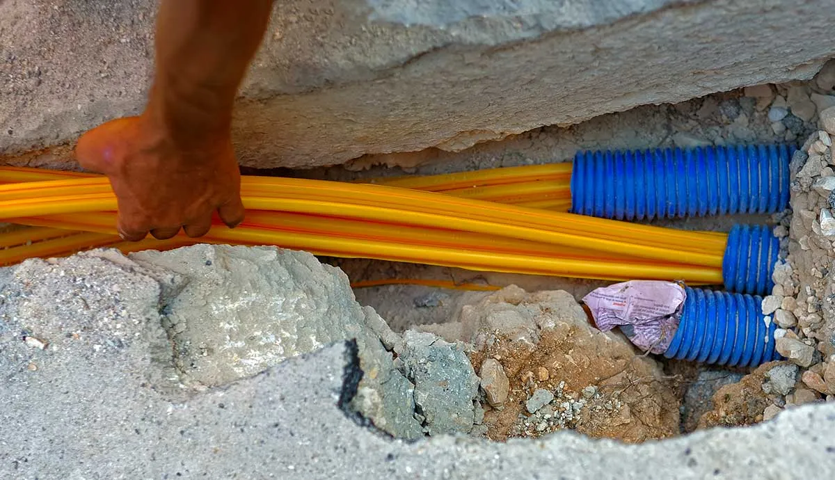 A worker's hand guides yellow cables into blue corrugated conduits in a concrete trench.