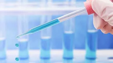 Gloved hand pipetting blue liquid into a test tube, with a rack of test tubes in the background.