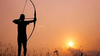 Silhouette of an archer, bow drawn, aiming in a field at sunset.