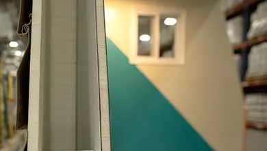 Close-up of a white siding panel’s cross-section, with a blurred background showing a diagonally painted teal and beige wall and a small window.