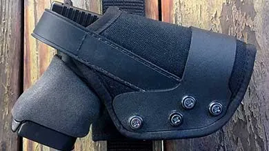 Close-up of a black handgun in a black holster with metal rivets, attached to a strap on a wooden surface.