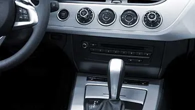 Close-up of a car interior showing the automatic gear shifter, silver dashboard with climate control knobs, and part of the steering wheel.