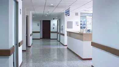 Empty hospital hallway with polished floors, white walls, and brown handrails, leading to a distant nurse's station.