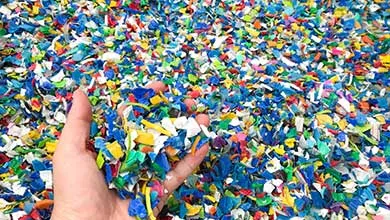 A hand holds colorful shredded plastic pieces above a large pile of similar plastic fragments.
