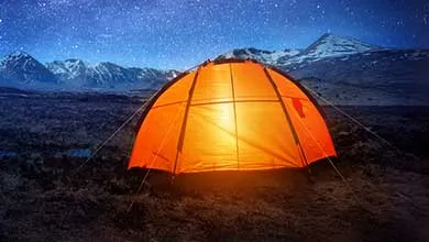 An orange dome tent glows warmly at night on dark ground, with snow-capped mountains and a starry sky in the background.