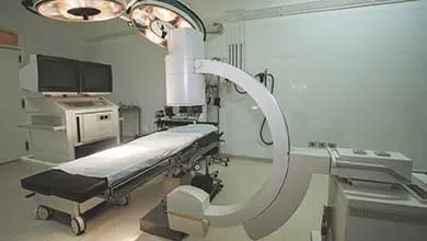 A clean medical procedure room with a large white C-arm X-ray machine positioned over a mobile operating table draped in white linens. Ceiling-mounted circular surgical lights illuminate the space, with medical monitors and diagnostic equipment along the white walls.