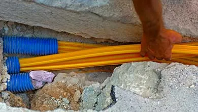 A worker's hand guides yellow cables into blue corrugated conduits in a concrete trench.