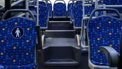 Empty bus interior, view down the aisle toward the back. Blue patterned seats; the nearest seat displays a white priority symbol showing a person with a cane.