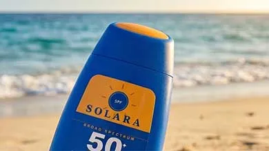 Close-up of a blue Solara SPF 50+ sunscreen bottle with an orange label, set against a blurred beach and ocean background.