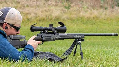 A person in a baseball cap and sunglasses lies on grass, aiming a sniper rifle mounted on a bipod.