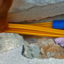A worker's hand guides yellow cables into blue corrugated conduits in a concrete trench.